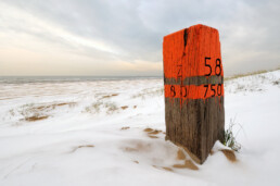 Houten strandpaal met oranje verf en gekerfde zwarte letters tussen besneeuwde, witte duinen op het Kennemerstrand bij IJmuiden.